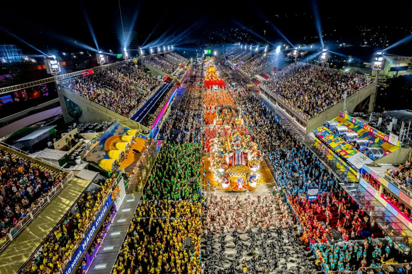 A samba school parading through the Sambadrome in Rio de Janeiro at night, a lit float flanked by thousands of costumed dancers and packed grandstands